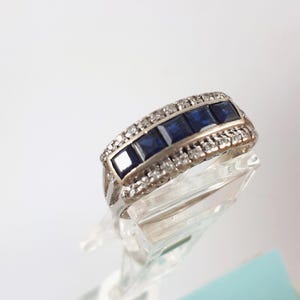 Può includere: Un anello d'argento con una fila di cinque pietre preziose quadrate blu scuro, affiancate da pietre più piccole e chiare. L'anello è esposto su uno sfondo bianco, evidenziando il suo design dettagliato. Un gioiello classico.