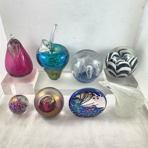 Pode incluir: Uma coleção de peças de arte em vidro coloridas, incluindo uma pera, uma maçã e várias esferas decorativas. As peças apresentam várias cores e designs, como redemoinhos, bolhas e padrões abstratos. Os objetos são exibidos em blocos de acrílico transparentes.