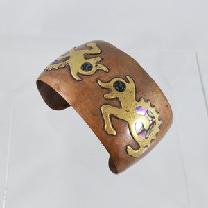 Op de afbeelding: Een koperen armband met een gouden ontwerp van gestileerde figuren. De figuren zijn ingelegd met iriserende abalone-schelp.