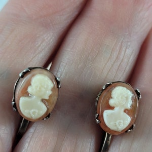 May include: A pair of vintage cameo earrings with a peach-coloured background and white cameo portraits. The earrings are silver-toned and have a clip-on closure.