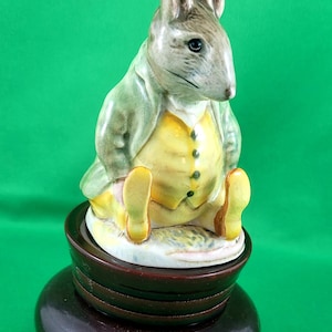 Può includere: Una statuetta in ceramica di un topo che indossa una giacca verde e un gilet giallo, seduto su una base di legno. Il topo ha un corpo marrone e un viso grigio.