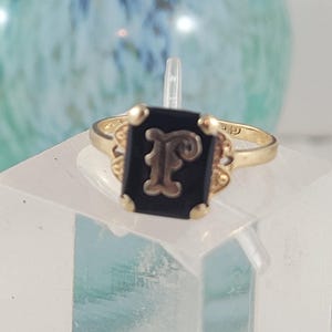 May include: A gold ring with a black onyx stone featuring a gold letter 'F' in the centre.