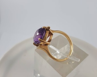 May include: A gold ring featuring a large, oval-shaped amethyst gemstone. The ring is displayed on a white stand.