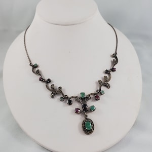 Puede incluir: Un collar de plata con un colgante de piedras preciosas verdes, rojas y negras. El colgante tiene forma de flor con una piedra central en forma de gota. El collar está hecho de una cadena delicada con pequeños detalles intrincados.
