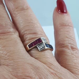 Peut inclure: Une bague en or avec un motif en chevron de pierres précieuses rouges et bleues. La bague est portée sur un doigt.