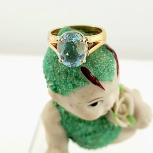 Könnte beinhalten: Ein goldener Ring mit einem großen, ovalen, hellblauen Edelstein. Der Ring befindet sich auf einer Figur mit grün strukturierten Haaren und Kleidung. Die Figur hat einen hellen Hautton und hält eine weiße Rose.