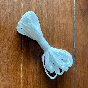 May include: A white elastic cord, folded in half, is shown on a wooden surface. The cord is approximately 1/4 inch wide.