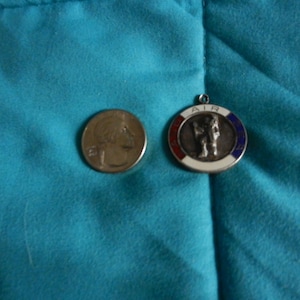 May include: A silver pendant with a figure in the center, surrounded by a red, white, and blue border. Next to it is a quarter coin. Both items are on a teal fabric surface.