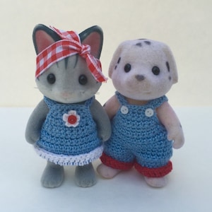 May include: Two small plush toys wearing crocheted overalls. The cat is gray with black ears and a red and white checkered headband. The dog is white with black spots and has a pink nose.