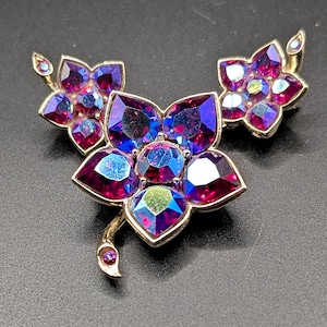 Peut inclure: Ensemble de bijoux vintage comprenant une broche et des boucles d'oreilles assorties. L'ensemble présente des motifs floraux avec des cristaux violets et bleus irisés sertis dans du métal doré. La broche a une tige et des détails de feuilles.