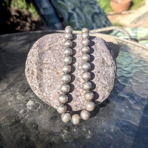 May include: A silver beaded necklace with round beads. The necklace is displayed on a gray stone, with a blurred background. The beads are strung together, creating a simple yet elegant design. The necklace is likely a piece of jewelry.