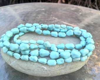 May include: A necklace composed of light blue, irregularly shaped beads. The beads are strung together, forming multiple strands that lay on a gray stone. The necklace is a light turquoise color, and the beads appear to be polished.