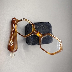 May include: A pair of folding lorgnette glasses with a brown tortoiseshell pattern and hexagonal frames. The handle and frame edges are decorated with small, clear embedded rhinestones arranged in floral and linear patterns.