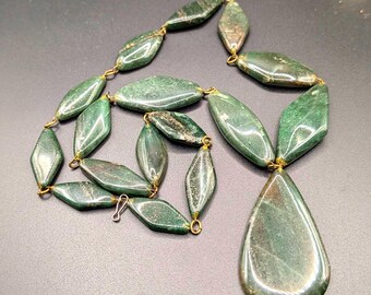 May include: A green gemstone necklace with a teardrop pendant. The necklace features oval and diamond-shaped beads connected by gold-colored links. The polished stones have a mottled appearance with variations in color.