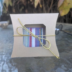 May include: A small, tan pillow box with a rectangular window showing a striped ribbon in purple, blue, and red. The box is tied with gold-coloured wire in a decorative bow. The background is blurred.