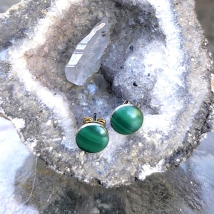 May include: A pair of round, green malachite stud earrings with silver posts. The earrings are set against a backdrop of a geode and a clear quartz crystal. The malachite has a distinctive banded pattern.