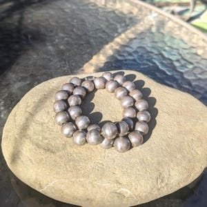 May include: A silver beaded necklace, composed of numerous round beads, rests on a smooth, light brown stone. The necklace is arranged in a circular shape, with the beads reflecting the sunlight. The background is blurred, suggesting an outdoor setting.