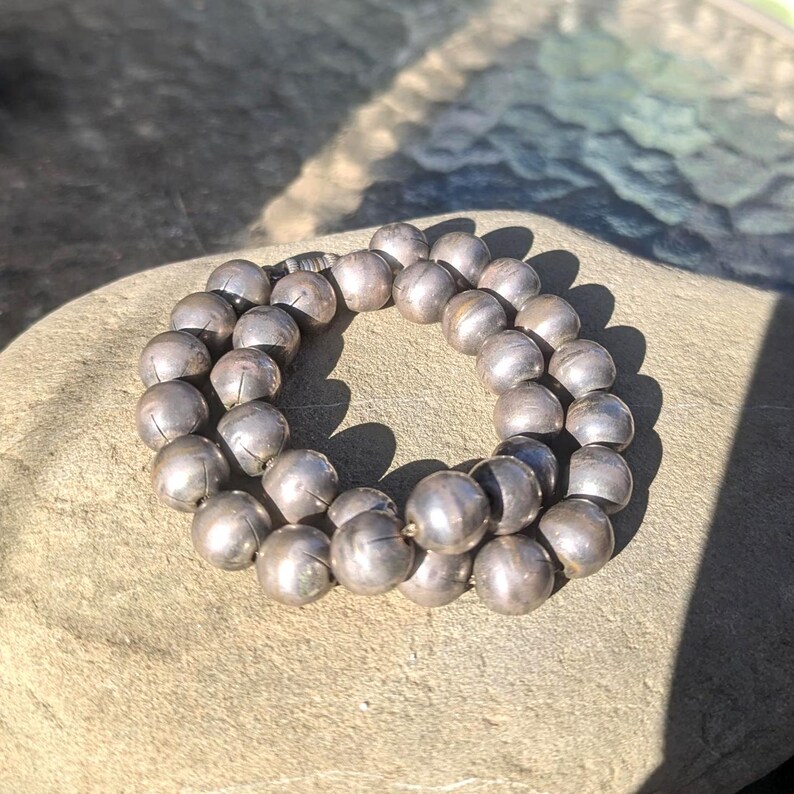 May include: A necklace composed of numerous silver-colored, spherical beads. The beads are arranged in a circular pattern, resting on a light brown surface. The beads have a slightly textured appearance, and the lighting creates shadows.