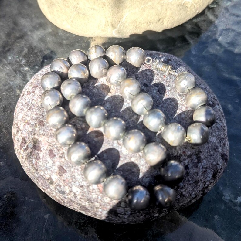 May include: A silver beaded necklace with a magnetic clasp, resting on a gray and brown stone. The beads are round and have a metallic sheen. The necklace is displayed outdoors in natural light.