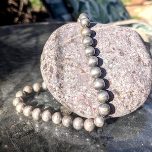 May include: A silver beaded necklace with large, round beads. The necklace is draped on a textured, gray stone, creating a contrast of textures and colors. The beads have a metallic sheen, reflecting light.