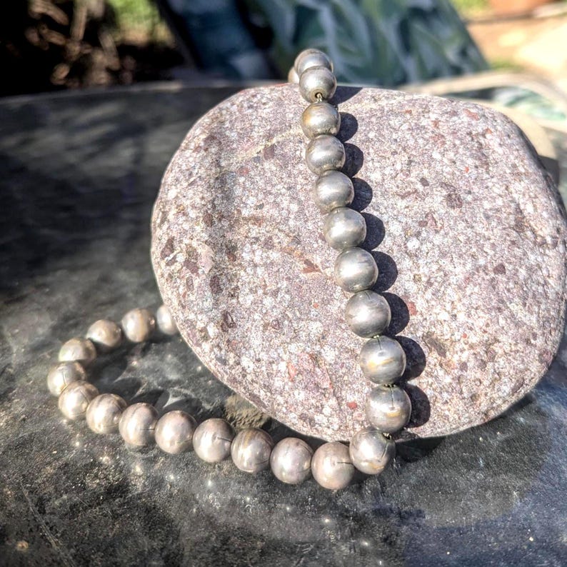 May include: A silver beaded necklace with large, round beads. The necklace is draped on a textured, gray stone, creating a contrast of textures and colors. The beads have a metallic sheen, reflecting light.