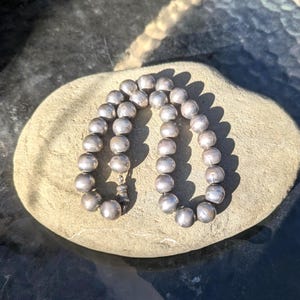 May include: A silver beaded necklace with a clasp, resting on a light brown stone. The necklace is composed of round, silver beads. The clasp is a detailed, decorative element. The beads have a slightly matte finish, reflecting the sunlight.