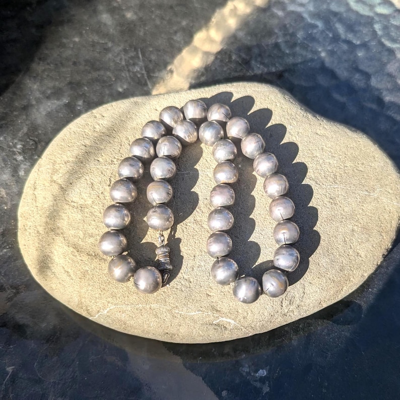 May include: A silver beaded necklace with a clasp, resting on a light brown stone. The necklace is composed of round, silver beads. The clasp is a detailed, decorative element. The beads have a slightly matte finish, reflecting the sunlight.