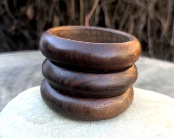 May include: Three dark brown wooden bangles stacked on top of each other. The bangles have a smooth, rounded design and a rich, natural wood grain. The bangles are displayed on a light-colored stone surface.