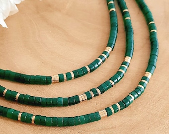 May include: Three strands of green beaded necklaces with gold-colored accents. The necklaces are displayed on a light brown wooden surface. A white flower is visible in the upper left corner.