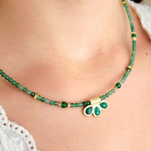 Peut inclure: Un collier délicat composé de petites perles vertes et d'éléments dorés. Le collier est orné d'un pendentif avec trois pierres vertes en forme de goutte d'eau dans un cadre doré. Le collier est porté.
