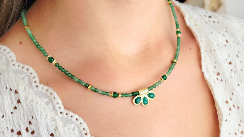 Peut inclure: Un collier délicat composé de petites perles vertes et d'éléments dorés. Le collier est orné d'un pendentif avec trois pierres vertes en forme de goutte d'eau dans un cadre doré. Le collier est porté.