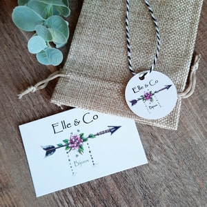 K&ouml;nnte beinhalten: Eine wei&szlig;e Karte mit dem Text "Elle & Co Bijoux" und einem stilisierten Pfeil mit einer rosa Blume. Die Karte liegt auf einer Holzoberfl&auml;che. Im Hintergrund befindet sich ein kleiner Jutesack mit einem wei&szlig;en und schwarzen Band. An dem Band ist ein rundes Etikett mit dem Text "Elle & Co Bijoux" befestigt.