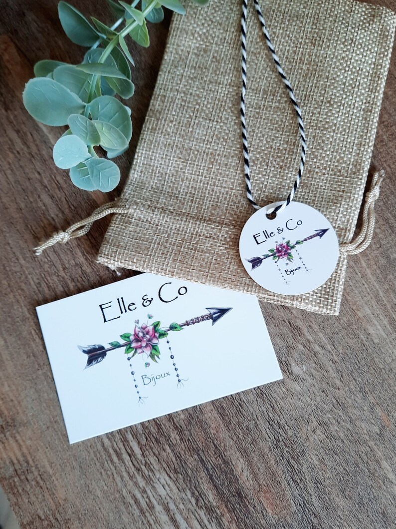 K&ouml;nnte beinhalten: Eine wei&szlig;e Karte mit dem Text "Elle & Co Bijoux" und einem stilisierten Pfeil mit einer rosa Blume. Die Karte liegt auf einer Holzoberfl&auml;che. Im Hintergrund befindet sich ein kleiner Jutesack mit einem wei&szlig;en und schwarzen Band. An dem Band ist ein rundes Etikett mit dem Text "Elle & Co Bijoux" befestigt.