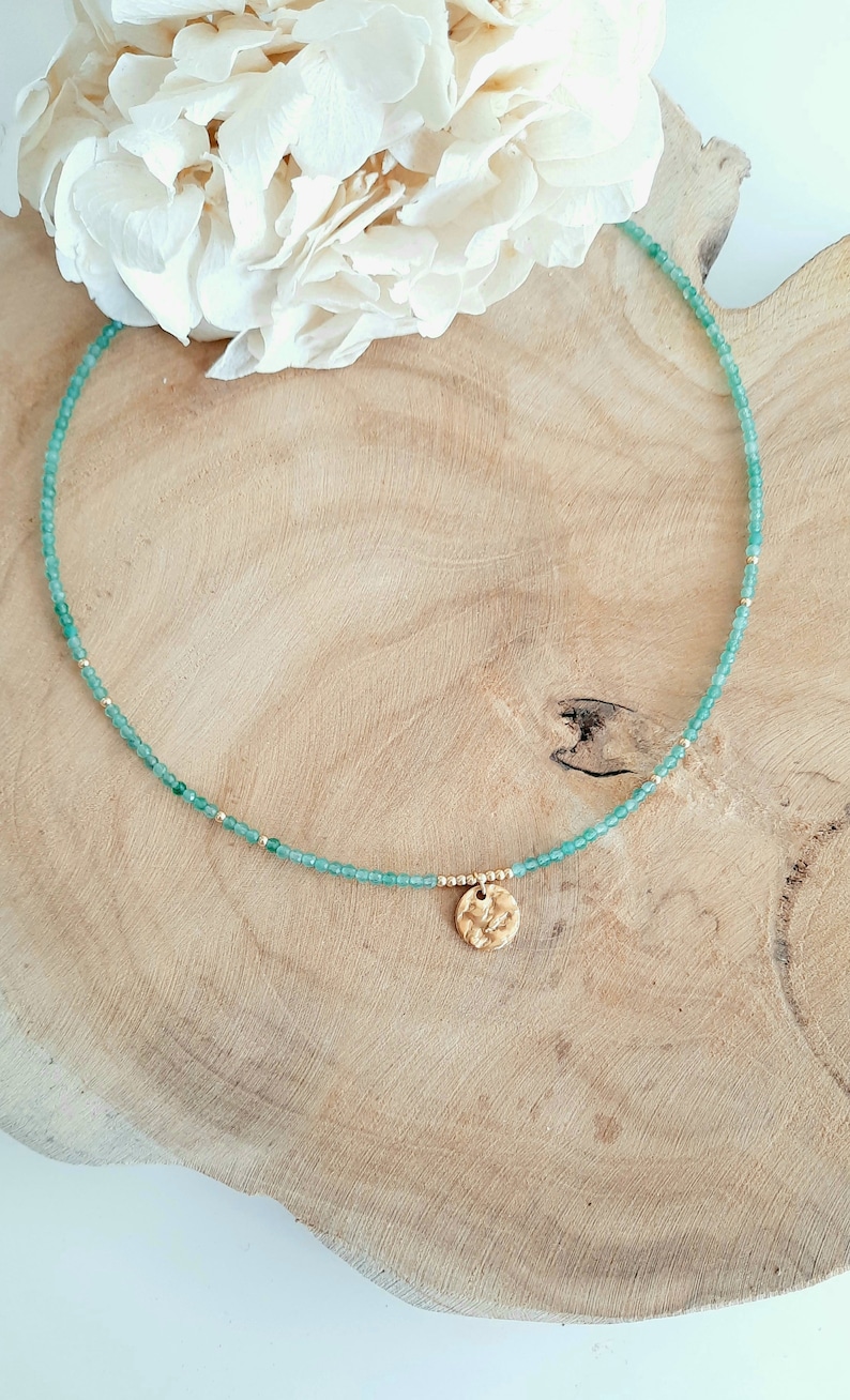 K&ouml;nnte beinhalten: Eine zarte gr&uuml;ne Perlenkette mit einem goldenen Anh&auml;nger. Die Kette liegt auf einer Holzoberfl&auml;che.