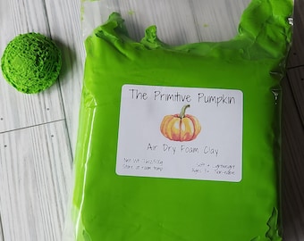 Puede incluir: Una bolsa de plástico transparente llena de arcilla de espuma verde brillante, junto a un pequeño objeto verde redondo y texturizado. La bolsa tiene una etiqueta que dice "The Primitive Pumpkin" y "Air Dry Foam Clay". El peso neto es de 500g.