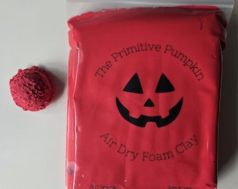 May include: A red air dry foam clay in a clear plastic bag labeled The Primitive Pumpkin with a black jack-o'-lantern graphic. The text reads 500 Grams, Soft & Lightweight, Store at Room Temp, and Ages 3+ Non-Edible. A small red clay sphere sits beside it.