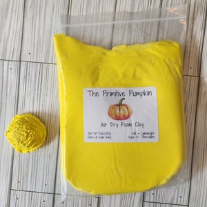 May include: A package of bright yellow air dry foam clay with a label that reads "The Primitive Pumpkin". A small, textured yellow clay shape is to the left of the package. The label also says "Net Wt 16oz/500g".