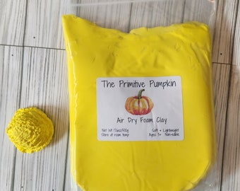 Puede incluir: Un paquete de arcilla de espuma de secado al aire de color amarillo brillante con una etiqueta que dice "The Primitive Pumpkin". A la izquierda del paquete hay una pequeña forma de arcilla amarilla texturizada. La etiqueta también dice "Peso neto 500g".