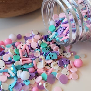 May include: A clear plastic jar filled with colorful sprinkles, including pastel pink, purple, blue, and green. The sprinkles are a mix of shapes, including circles, stars, and hearts. The jar is tipped over, spilling the sprinkles onto a white surface.