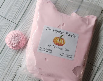 Puede incluir: Un paquete de arcilla de espuma rosa que se seca al aire de "The Primitive Pumpkin" con una pequeña bola rosa texturizada. La etiqueta dice "Air Dry Foam Clay" y "Peso neto 500g". El producto es suave, ligero y no comestible.