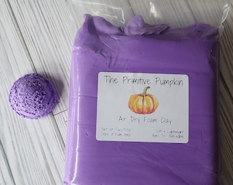 Puede incluir: Un paquete de arcilla de espuma de secado al aire púrpura de "The Primitive Pumpkin" con una pequeña bola púrpura texturizada. La etiqueta dice "Air Dry Foam Clay" y "Peso neto 500g". La arcilla es suave, ligera y no comestible.