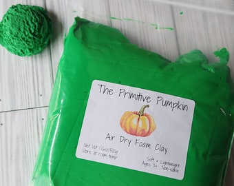 Puede incluir: Una bolsa de plástico transparente llena de arcilla de espuma verde brillante, junto a una pequeña bola de arcilla verde. La etiqueta dice "The Primitive Pumpkin" con una ilustración de calabaza. El texto también incluye "Air Dry Foam Clay", "Peso neto 500g" y "Ages 3+".