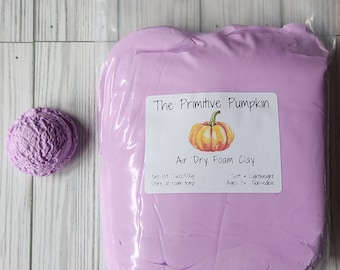 Puede incluir: Un paquete de arcilla de espuma de secado al aire de color lila claro de "The Primitive Pumpkin" con una pequeña bola texturizada del mismo color. La etiqueta del paquete incluye un gráfico de calabaza y el texto: "Air Dry Foam Clay". El peso neto es de 500g.