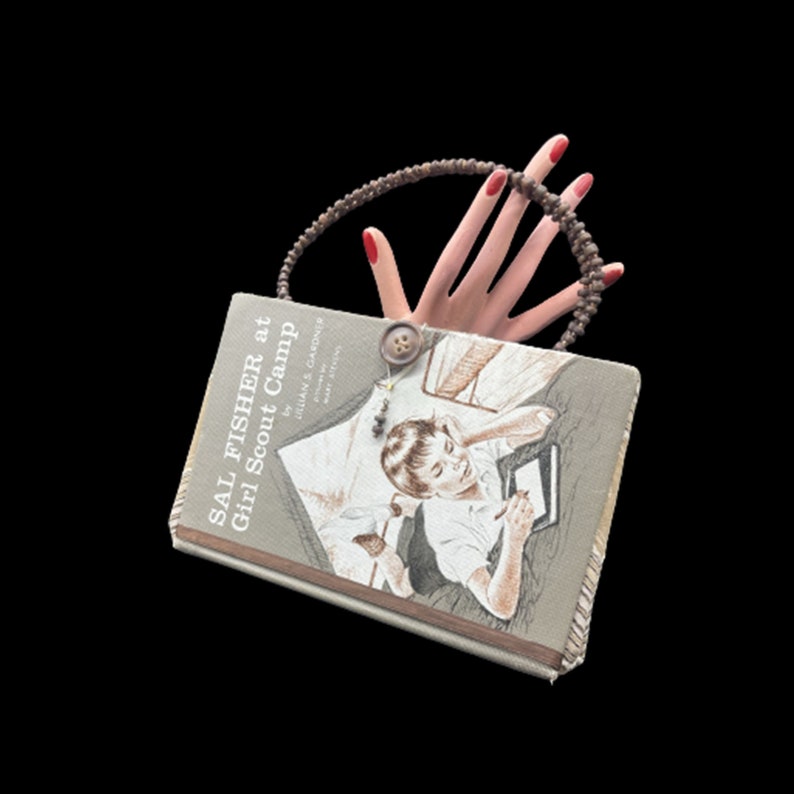 May include: A vintage book titled "Sal Fisher at Girl Scout Camp" by Dorothy Gardiner with a brown beaded necklace and a hand with red nail polish on the cover.