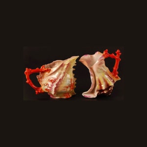 May include: Two porcelain creamers shaped like seashells with red coral handles. The creamers are decorated with pink, yellow, and white glaze.