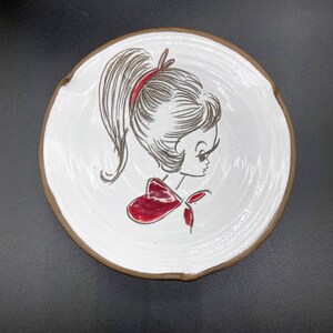 May include: A white ceramic ashtray with a brown rim features a black and white line drawing of a girl with a red bow in her hair. The girl is looking to the right.