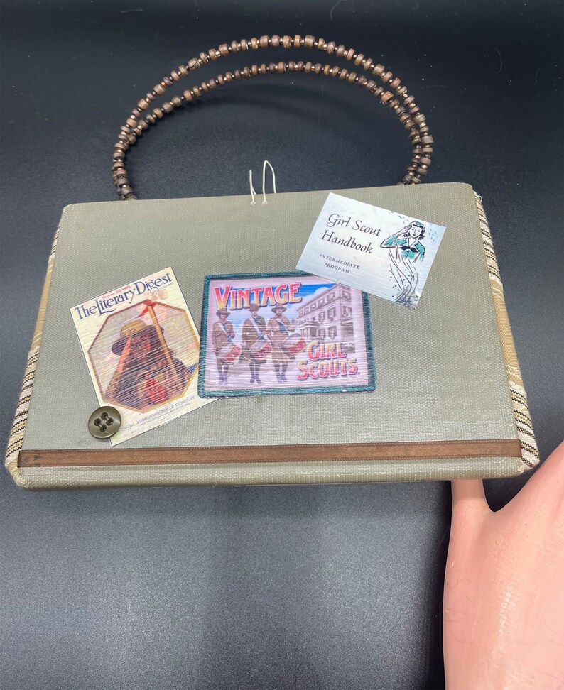 May include: A green fabric purse with a wooden beaded handle. The purse has a vintage Girl Scout patch, a "The Literary Digest" magazine cover, and a Girl Scout Handbook.