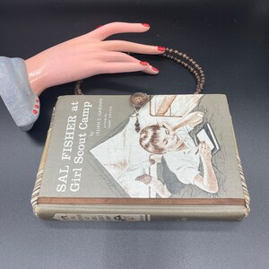 May include: A vintage book titled "Sal Fisher at Girl Scout Camp" by Lillian S. Gardner, illustrated by Mary Stevens. The book is brown with a plaid pattern on the spine and a picture of a girl writing in a notebook.