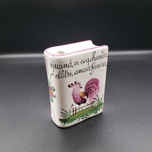 May include: A white ceramic book-shaped trinket with a pink rooster illustration and the French text "Quand ce coq chantera Notre amour finira".