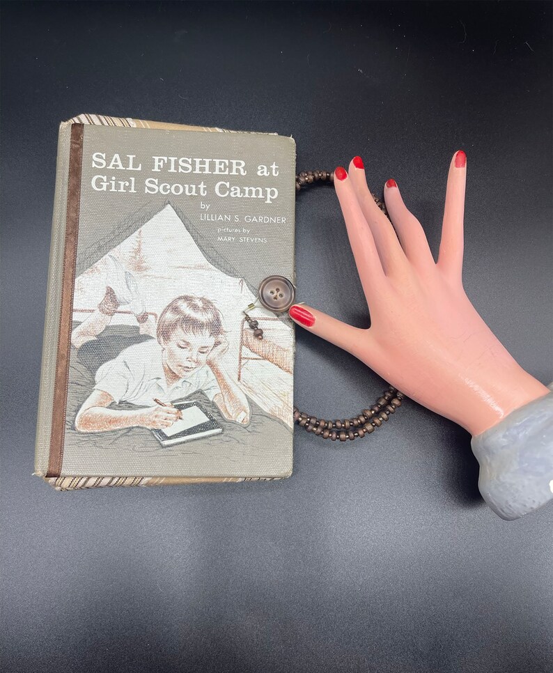 May include: A vintage book titled "Sal Fisher at Girl Scout Camp" by Lillian S. Gardner with illustrations by Mary Stevens. The book cover features a drawing of a girl sitting in a tent, writing in a notebook.